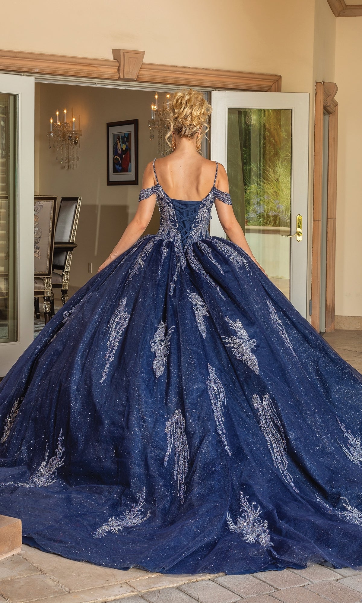 A woman wearing the Quinceanera Dress 1732 By Dancing Queen, an off-the-shoulder navy blue A-line ball gown with silver lace embroidery, stands with her back to the camera holding out the skirt in a room with open glass doors.